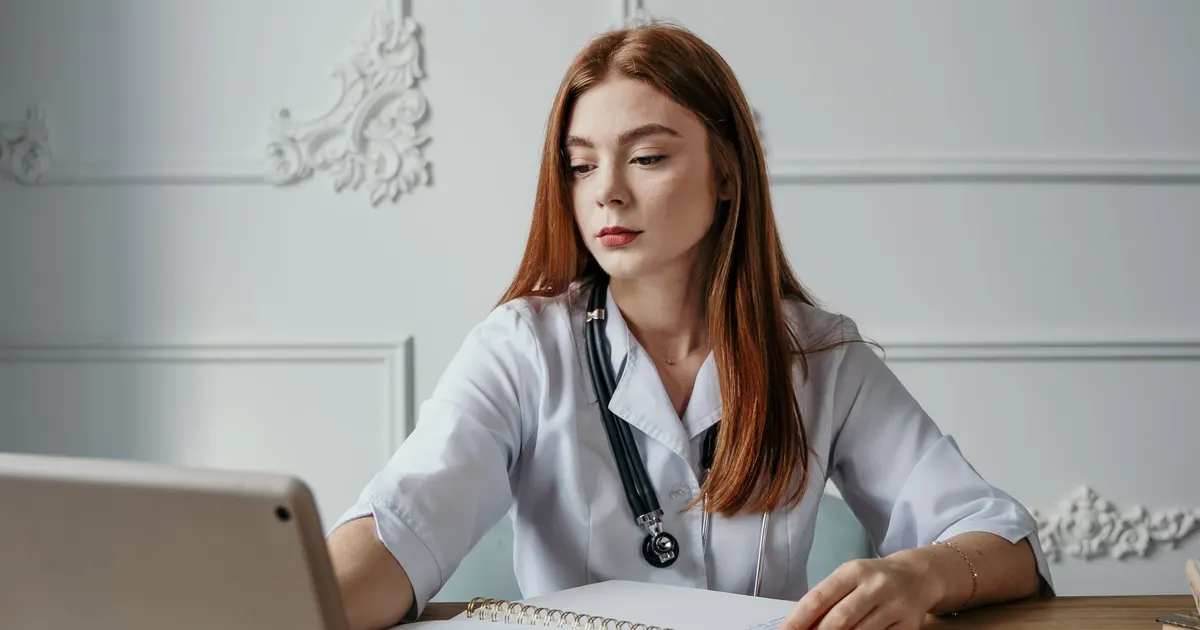 Doctora con estetoscopio trabajando en un computador portátil sobre un escritorio con cuadernos y medicamentos
