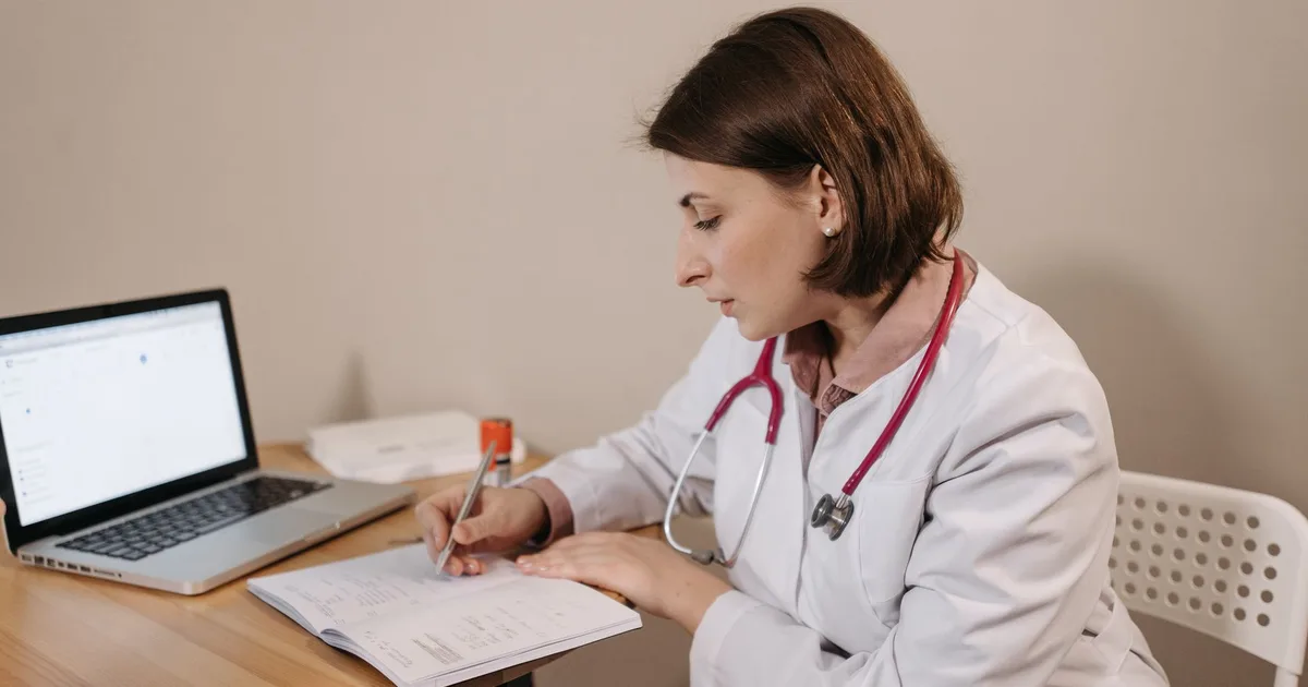 Una doctora con bata de laboratorio revisa información en un laptop en su consultorio