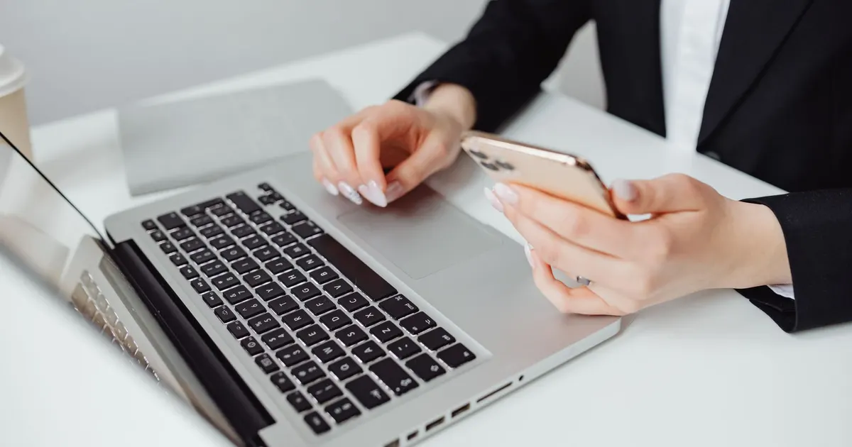 Mujer con ropa formal usando un laptop y un teléfono celular sobre un escritorio de oficina.