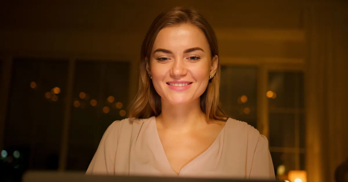 Mujer sonriente usa su laptop de noche en un ambiente interior cálido y acogedor.