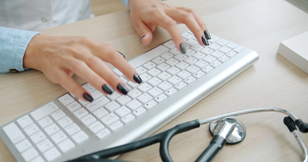 Primer plano de un profesional de salud escribiendo en un computador de escritorio, con un estetoscopio sobre la mesa.
