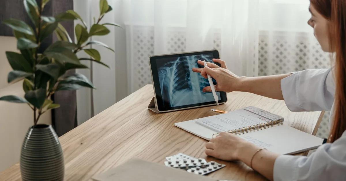 Profesional de salud examina una imagen de rayos X en una tablet junto a notas médicas en un escritorio