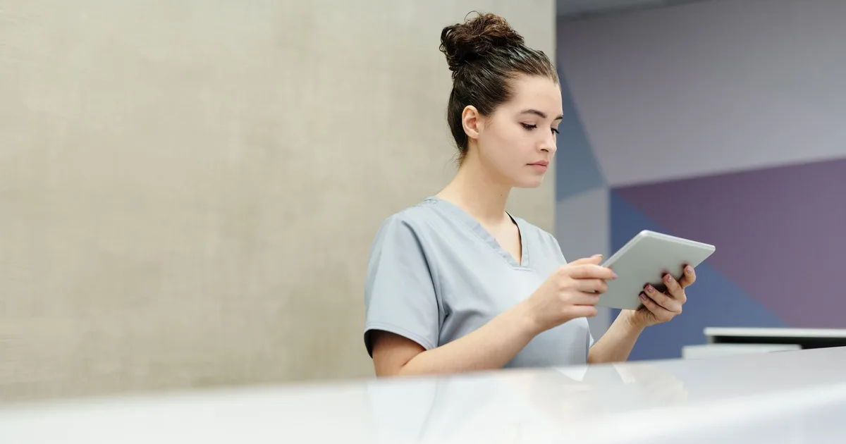 Recepcionista en scrubs usando una tablet en el mostrador de recepción de una clínica