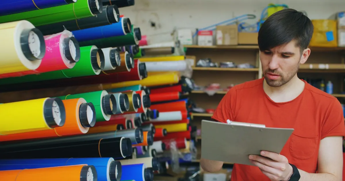 Hombre en una tienda revisa con una tablilla el inventario de rollos de papel de colores en una estantería