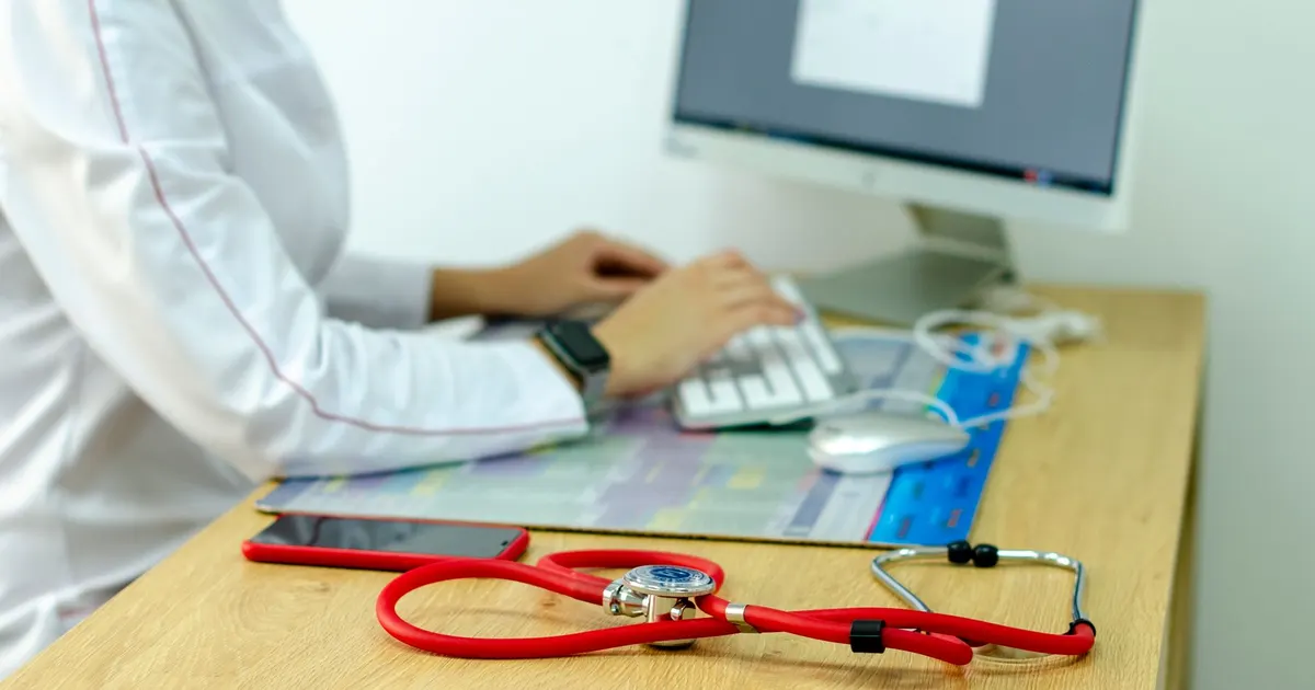 Profesional de salud escribiendo en un computador de escritorio, con un estetoscopio apoyado junto al teclado.